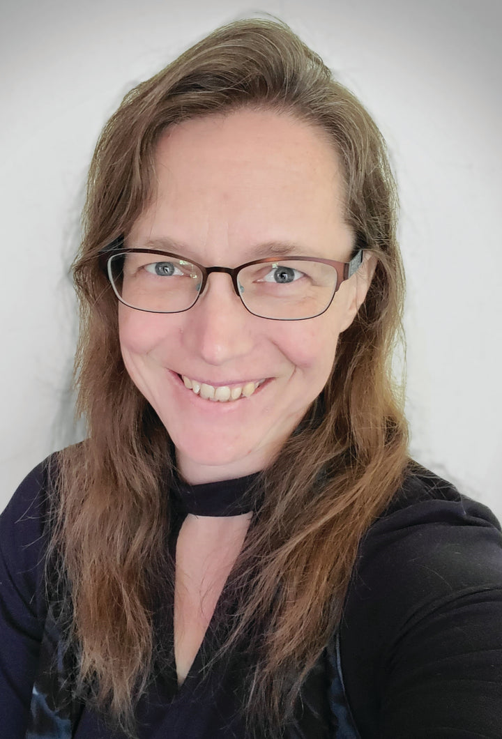 A woman with long brown hair and glasses smiles warmly at the camera in a black top against a light background, reflecting her passion for personal growth and the Guardian Tarot by Schiffer Publishing, Ltd.