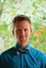A man with short light brown hair in a bright blue shirt and plaid bow tie stands outside with greenery, smiling slightly while holding The Witch’s Oracle, 2nd Edition by Schiffer Publishing, Ltd.