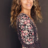 A woman with long, wavy, light brown hair stands sideways, smiling in emotional well-being. She wears a black floral dress. "Listening to Flowers" by Schiffer Publishing, Ltd. is set against a dark, plain background.