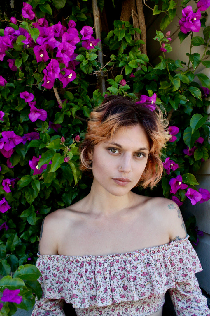 Radiating emotional well-being, a person with short, wavy, light brown hair and a floral top stands before green leaves and magenta flowers, inspired by "Listening to Flowers" from Schiffer Publishing, Ltd.