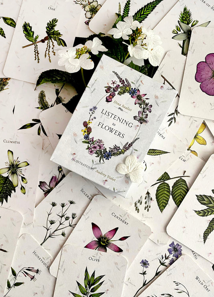 The book "Listening to Flowers" by Schiffer Publishing, Ltd. rests on illustrated flower cards featuring botanical drawings and Bach flower essence insights, surrounded by white blooms and greenery.