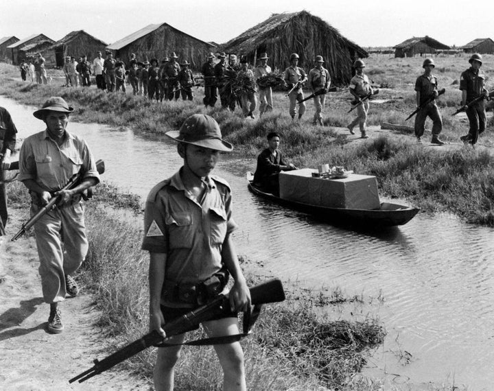 Armed soldiers patrol a canal beside a man paddling a boat with thatched huts behind them in rural South Vietnam during the Vietnam War, where "The Fighting Fathers" by Schiffer Publishing, Ltd. highlights their covert activities.