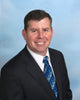 A man in a black suit, white shirt, and blue patterned tie smiles at the camera against a light blue gradient background, echoing the sleek professionalism featured in "Boeing 777" by Schiffer Publishing, Ltd.