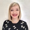 A smiling person with straight blonde hair, red lipstick, and blue eyes stands in front of a light background wearing a black sweater adorned with white bow patterns—getting ready to begin their moon journaling session using the Nocturnal Garden Lenormand deck by Schiffer Publishing, Ltd.
