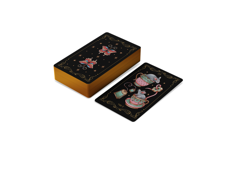 A stack of Nocturnal Garden Lenormand cards by Schiffer Publishing, Ltd. is shown with gold edges. One card displays a teapot, cup, and heart-shaped steam in whimsical art; the card backs have celestial and floral designs on black.