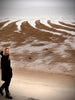 A person in a black coat smiles on a sandy beach with tidal patterns, enjoying Cape Cod Cones by Schiffer Publishing, Ltd., as clouds linger over the calm sea.