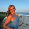 A young woman stands on a sandy beach at sunset, smiling warmly with her hands in her pockets, surrounded by nature patterns inspired by the Wild & Whimsical Woodland Coloring Book from Schiffer Publishing, Ltd.