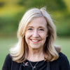 A smiling middle-aged woman with long blonde hair, in a black top and necklace, stands outdoors with a blurred green background, perhaps getting inspired to use Schiffer Publishing, Ltd.’s “Designing for Foundation Paper Piecing.”.