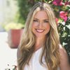 A woman with long blonde hair and a white sleeveless top smiles outdoors near pink flowers, holding Schiffer Publishing, Ltd.'s Watercolor the Easy Way Paint-Your-Own Watercolor Cards Autumn kit in soft sunlight.