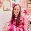 A woman with long brown hair, wearing a pink striped sweater, sits smiling in a colorful room decorated with flowers, motivational art, and adorable stickers from Schiffer Publishing, Ltd.’s *A Day in Cozy Town: The Cute Café Coloring and Sticker Book*.