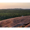 Two people sit on a large, rocky outcrop overlooking the Texas Hill Country at sunset.