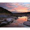 A scenic Texas sunset from Texas Hill Country features pink and orange clouds reflecting in the calm water of Pedernales Falls State Park, flowing past rocky river banks.