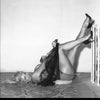 A black and white photo from Schiffer Publishing, Ltd.’s "Striptease Artists of the 1950s" shows a smiling woman in lingerie and heels, lying on her back with legs up on a railing, capturing classic burlesque style against a plain background.