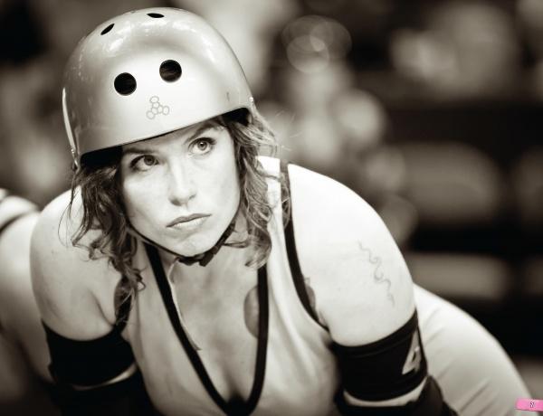 A curly-haired roller derby skater in protective gear looks ahead with intensity. "No Mercy" by Schiffer Publishing, Ltd. captures raw determination and focus in black-and-white, showcasing the heat of modern roller derby action.