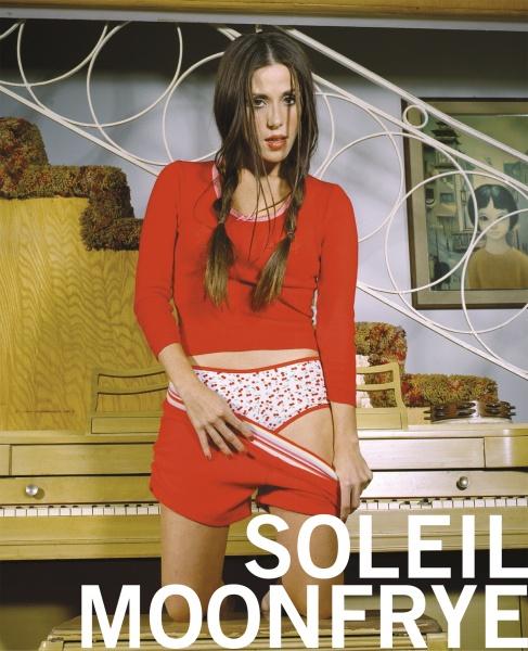 A woman with long brown hair wearing a red crop top and shorts stands by a piano, pulling down her shorts to reveal star-patterned underwear. Large text reads The Worst of While You Were Sleeping by Schiffer Publishing, Ltd., echoing a pop culture book cover.