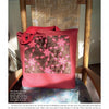 A red tote bag made with mad weave, featuring a brown and pink geometric Dancing Star pattern, from Hex Weave & Mad Weave by Schiffer Publishing, Ltd., sits on a chair with a woven backrest in sunlight.