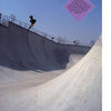 A skateboarder does a trick on the curved edge of a large Skateboard Parks facility by Schiffer Publishing, Ltd., under clear skies. Shadows stretch across the smooth concrete and a tall fence lines the park’s top edge.