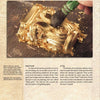 A hand uses a brush to apply gold leaf onto an ornate raised surface, with loose gold flakes nearby. The textured background shows text about the history of gold and gilding techniques from "GOLD" by Schiffer Publishing, Ltd.