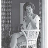 A woman sits on a wicker chair in classic 1950s style, smoking and gazing at the camera. This black-and-white image captures the allure featured in "Femmes Fatales of the 1950s" by Schiffer Publishing, Ltd. Patterned curtains and art set the scene.