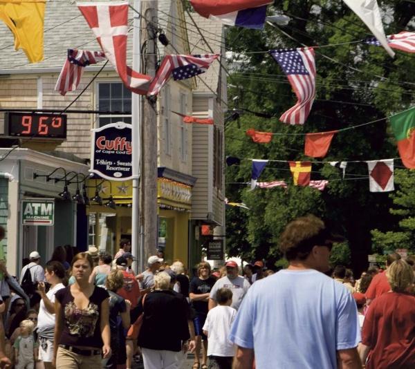 A lively street scene in Provincetown, MA, with people strolling beneath colorful flags and a sign reading 96°F. Shops and trees line the sunlit avenue—just one of many captivating images in "Provincetown Perspectives" by Schiffer Publishing, Ltd.