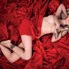A tattooed blonde woman poses on red satin sheets in a vintage style, inspired by "Queens of Hearts" from Schiffer Publishing, Ltd., with one arm across her chest, the other on her shoulder, gazing up at the camera.