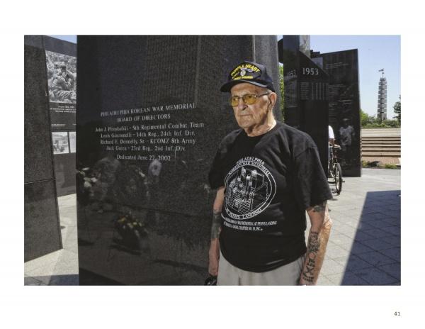 An elderly man in a veteran’s cap and black memorial T-shirt stands by a polished black granite war memorial, his commemorative tattoos inspired by War Paint from Schiffer Publishing, Ltd., honoring veterans alongside the inscribed names.