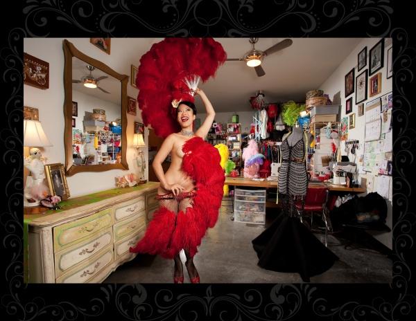 A beaming burlesque star in a red feathered outfit strikes a pose in a vivid dressing room—a perfect photo from *It's All That Glitters* by Schiffer Publishing, Ltd., ideal for fans of glamour and burlesque photography.