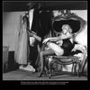A woman in vintage lingerie adjusts her stockings before a mirror, surrounded by 1950s costumes—a scene echoing the glamorous burlesque of "Striptease Artists of the 1950s" from Schiffer Publishing, Ltd.
