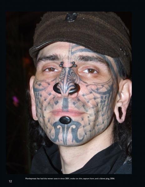 Featured in "Ink & Steel" by Schiffer Publishing, Ltd., this portrait captures a person with extensive facial tattoos, piercings (septum horn), chin implants, stretched earlobes, and a brown cap, facing the camera in dark clothing.